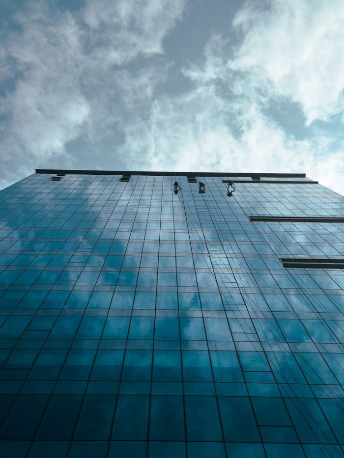 Three window cleaners working on a tall glass skyscraper against a cloudy sky.